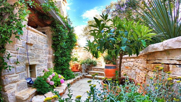 A stone-walled courtyard with a variety of plants and flowers, a small tree, and a rustic stone bench. - Villa in Murter Near Kornati National Park (Murter-Kornati)