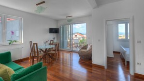 A spacious living area with a wooden floor, a green sofa, a dining table with chairs, and a view of the outdoors through large windows. - Villa in Murter Near Kornati National Park (Murter-Kornati)