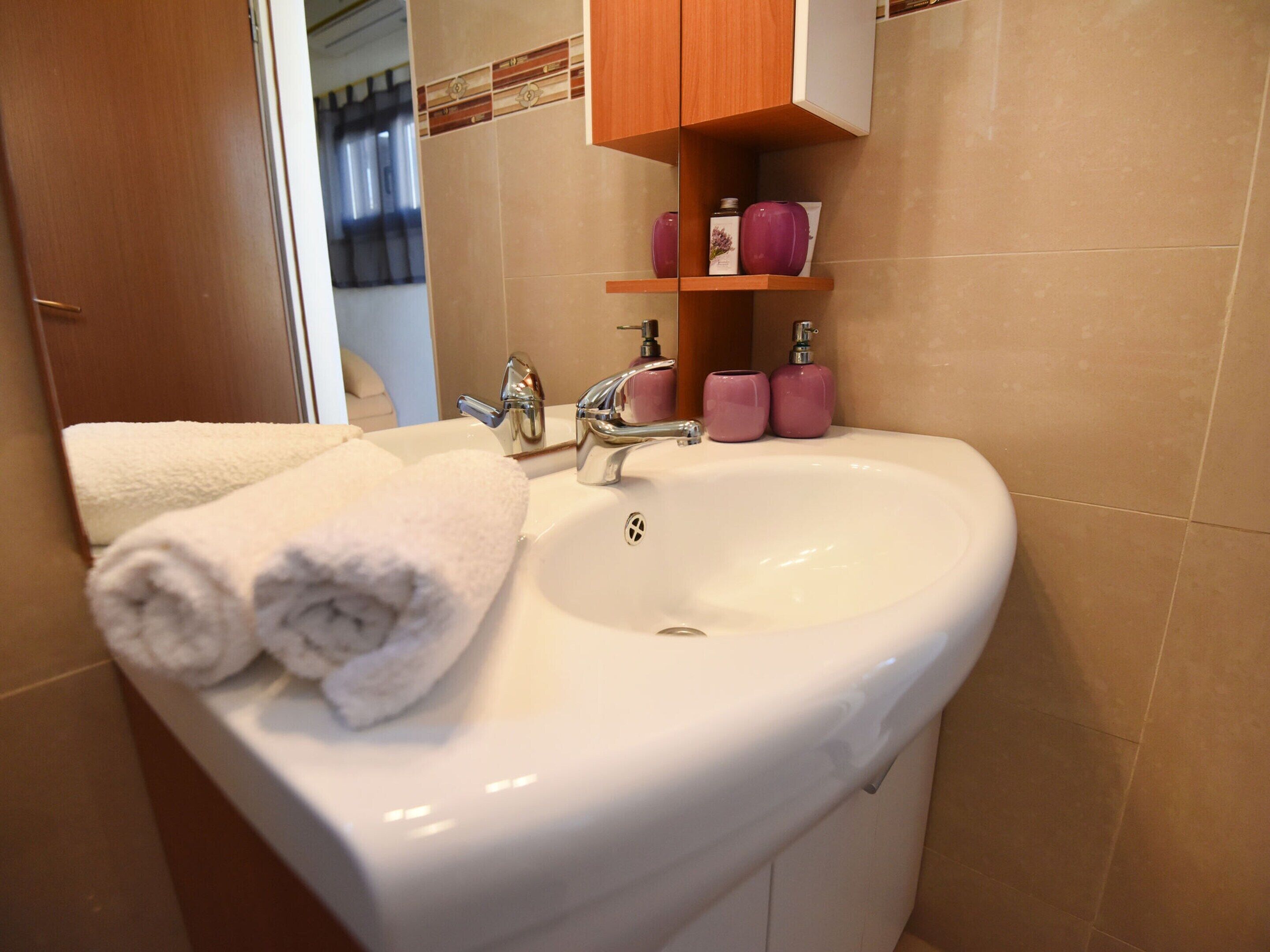 Une salle de bain avec un lavabo blanc, deux robinets et un miroir au-dessus. On peut voir deux bouteilles roses sur une étagère au-dessus du lavabo.