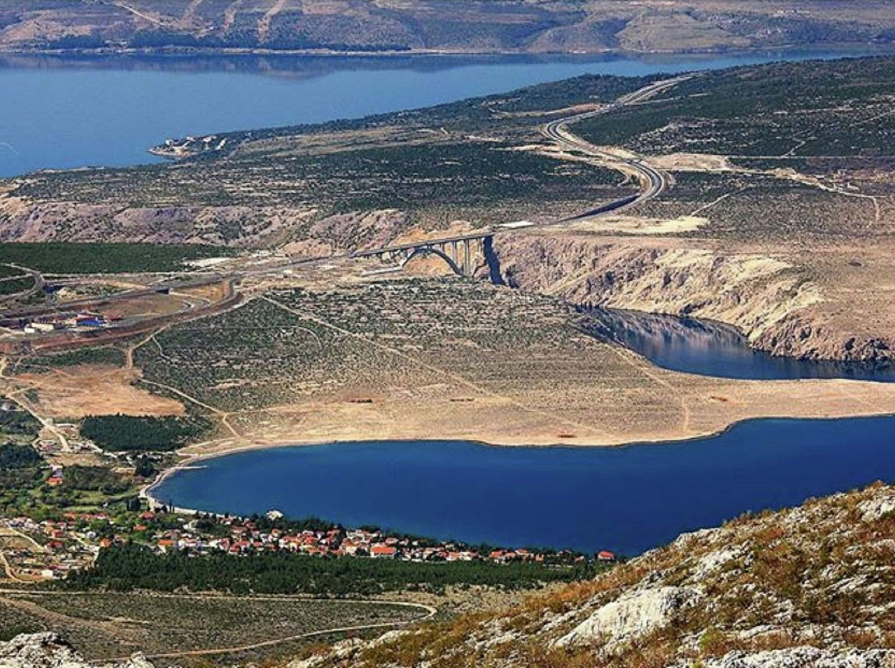 Paesaggio costiero con un ponte, un'ampia distesa d'acqua e un piccolo insediamento con edifici dai tetti rossi.
