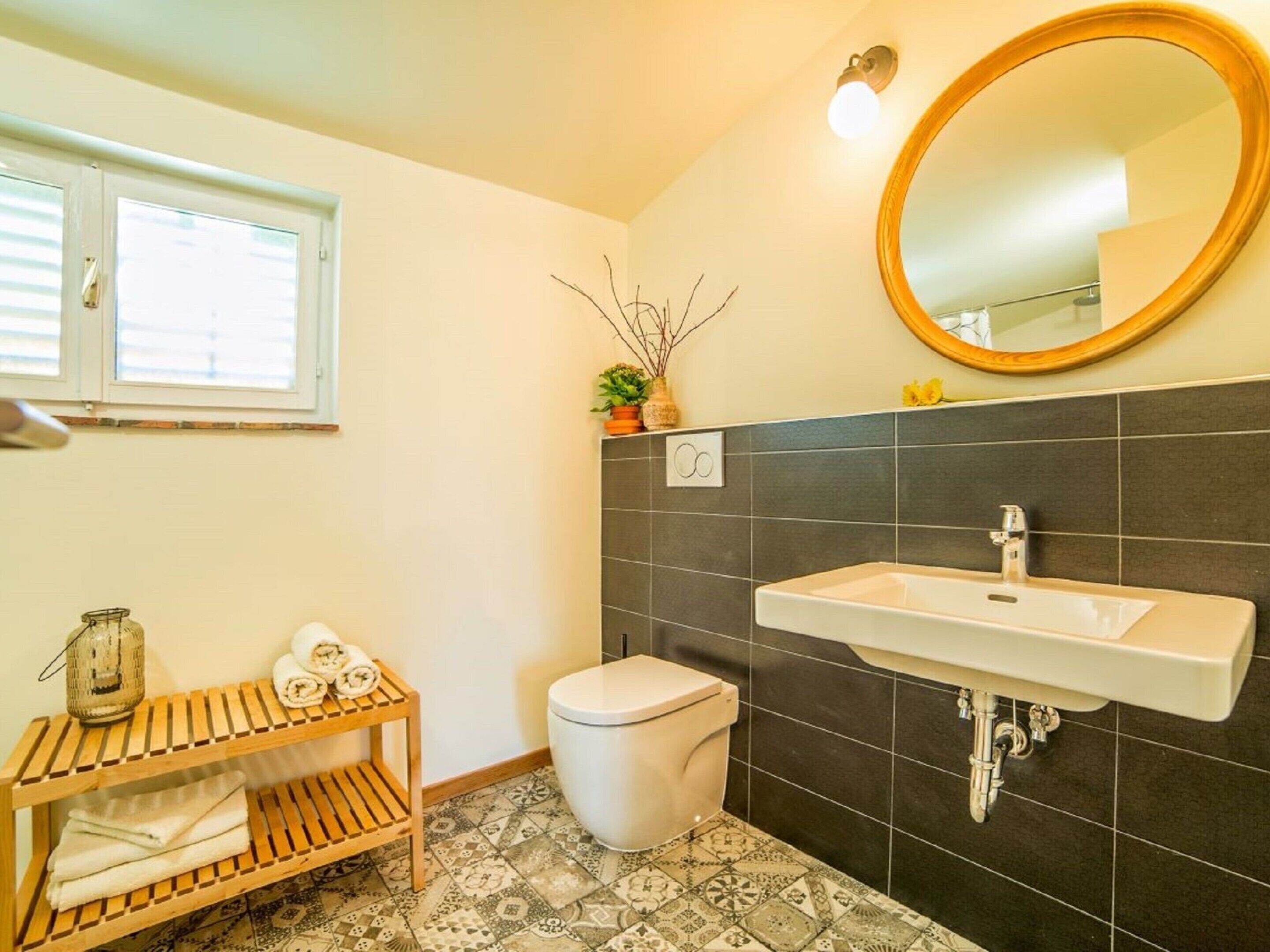 A bathroom with a white sink, dark tiled walls, a toilet, a wooden shelf with towels, a potted plant, and a round mirror.