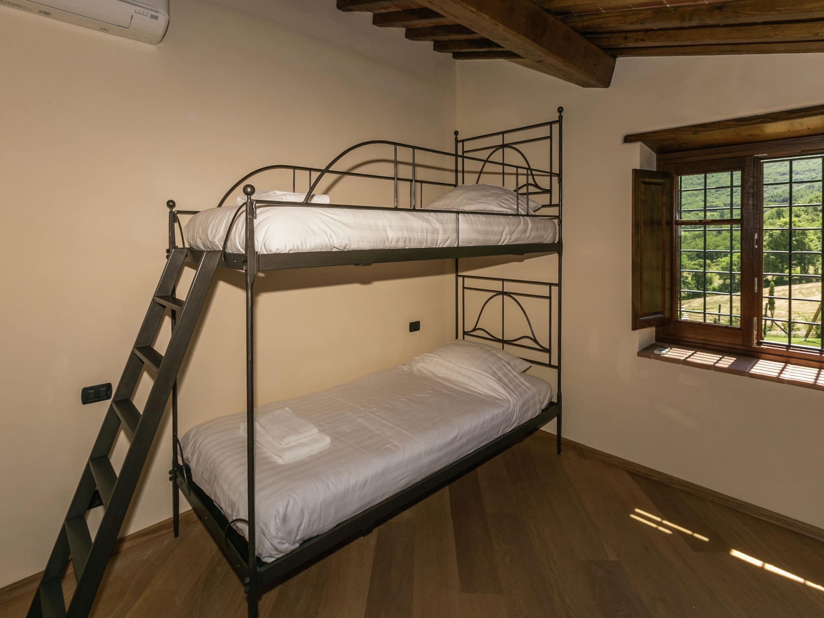 A bunk bed with a ladder, a window with a view of greenery, and a wooden ceiling.