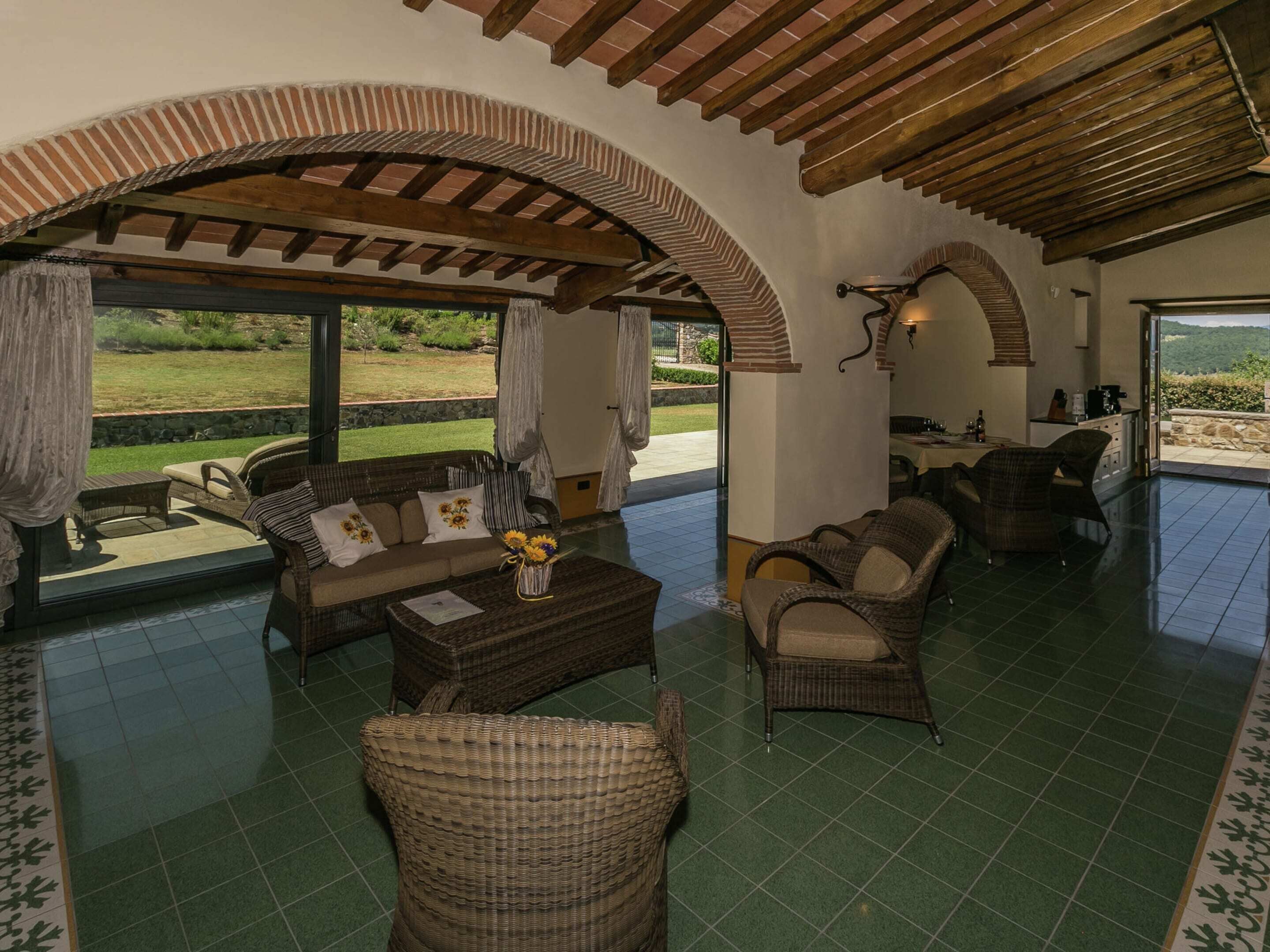 Amplia sala de estar con vista al jardín, amoblada con sillas de mimbre y un sofá, y una cocina visible a través de una puerta abierta.