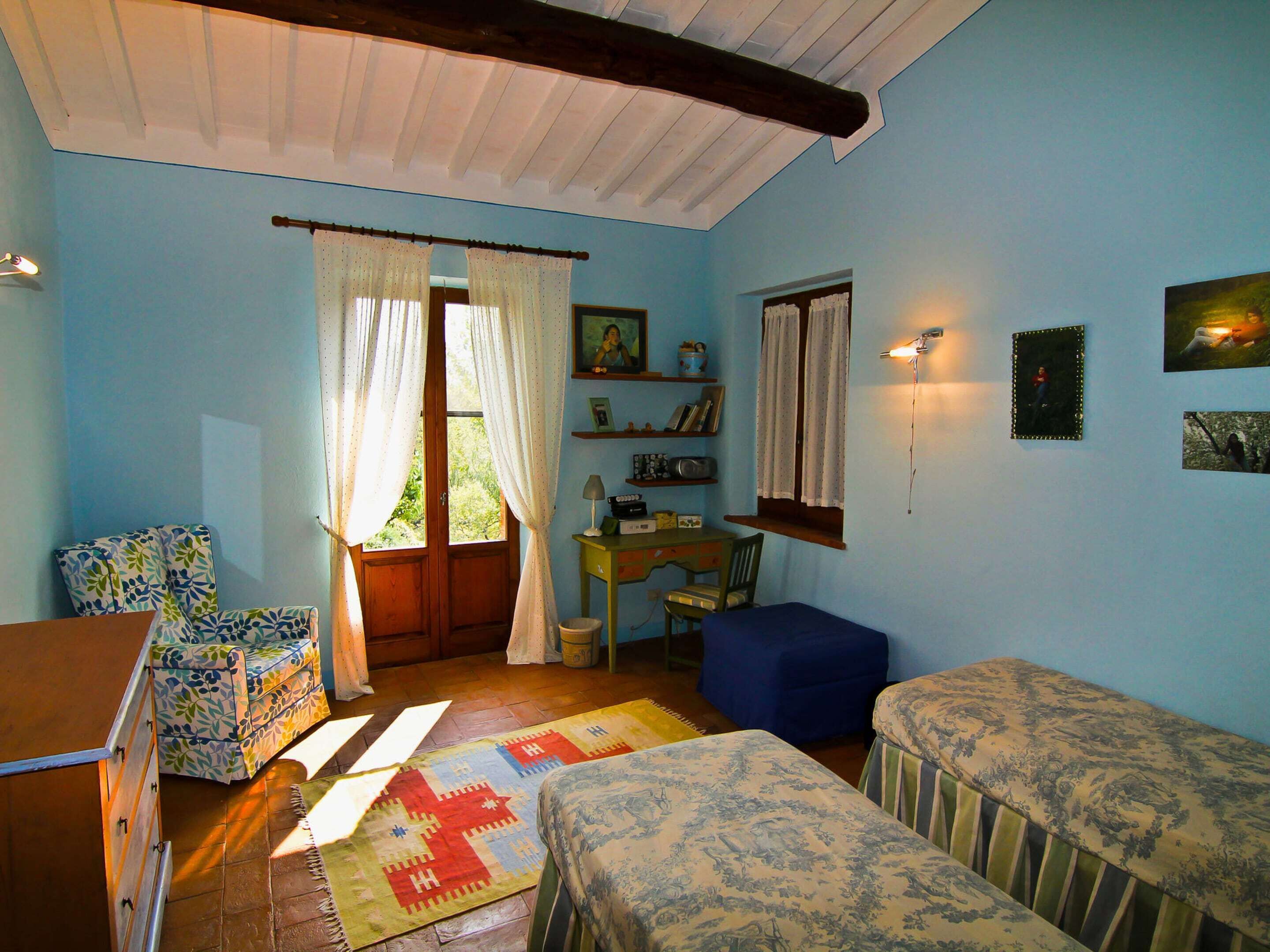 A room with a wooden ceiling, a desk with a chair, two beds, a window with curtains, and wall-mounted lights.