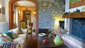 A cozy living room with a stone wall, a fireplace, a dining area with chairs, and a sofa with patterned cushions.