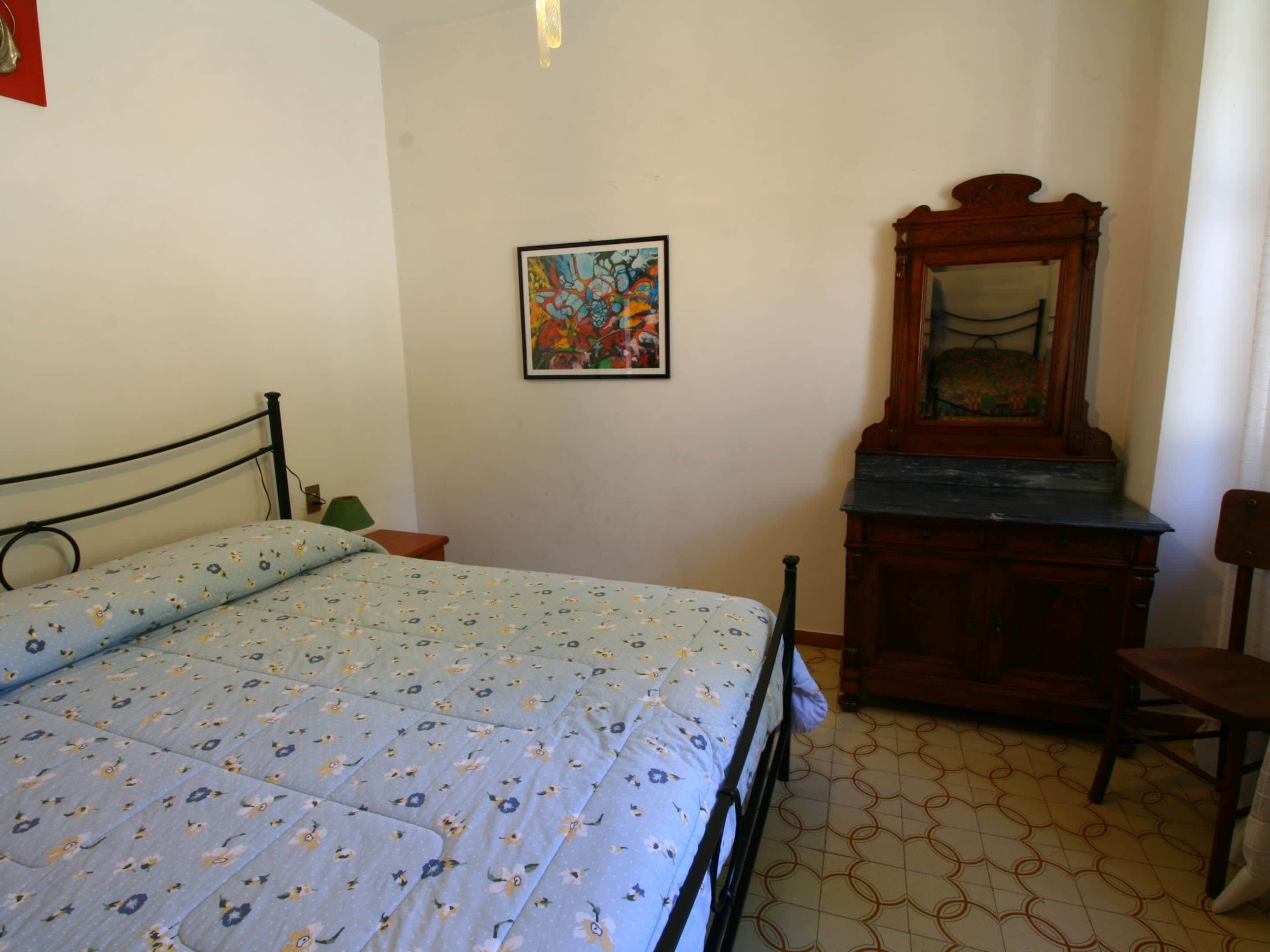 Ein Zimmer mit einem Bett, einem Schminktisch, einem Stuhl, einem Bild und einem Spiegel.