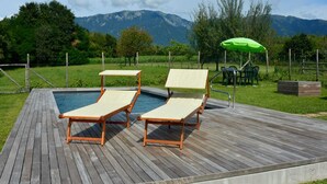 Un pontile in legno con due poltrone da relax e un ombrellone verde, con vista su una piscina e montagne.