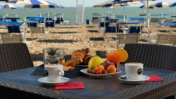 Mesa de desayuno junto a la playa con croissants, fruta y café, con vistas a la playa con sombrillas y sillas de descanso.