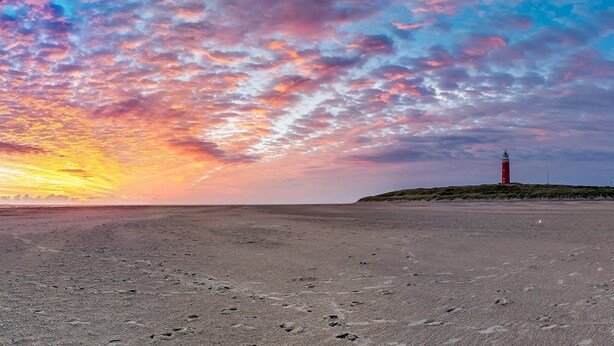 Uma praia com um farol numa pequena colina, sob um céu colorido.