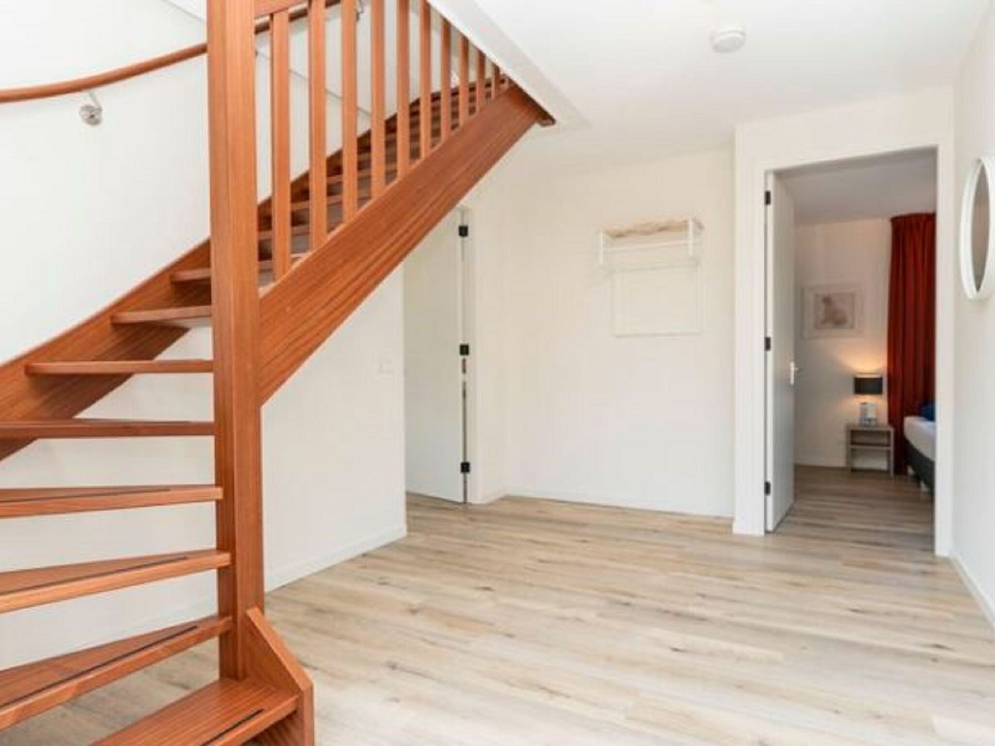 Un interior moderno con una escalera de madera, suelo de madera clara y un pasillo que lleva a un dormitorio con cama y mesita de noche.