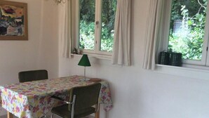 Une pièce avec une table recouverte d’une nappe à motifs floraux, une chaise verte, une lampe et une fenêtre avec des rideaux.