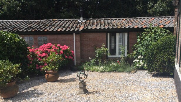 Une cour avec un sol de gravier, un bâtiment en briques surmonté d’un toit en tuiles, et des plantes en pot ornées de fleurs roses.