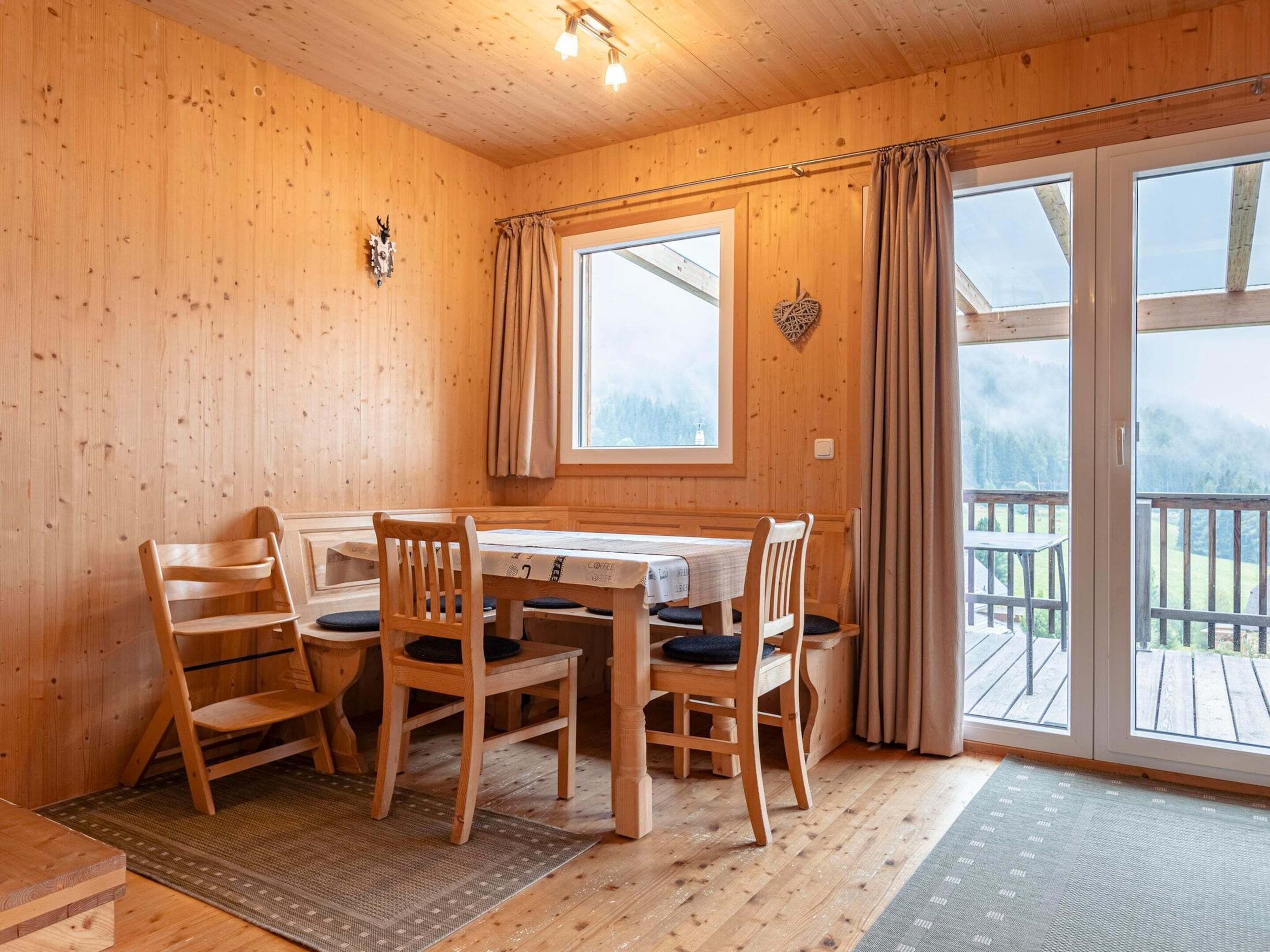 A wooden cabin with a dining area featuring a table and chairs, a large window with curtains, and a view of a deck and forested landscape.
