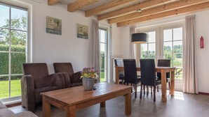 Ein geräumiger Wohnbereich mit einem Holztisch, zwei Sofas und einem Esstisch mit Stühlen. Große Fenster sorgen für natürliches Licht und Ausblicke ins Freie.