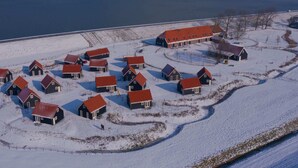 Schneebedeckte Landschaft mit mehreren Häusern und einem großen Bauernhaus, wahrscheinlich ein Bauernhof, mit einem Gewässer im Hintergrund.