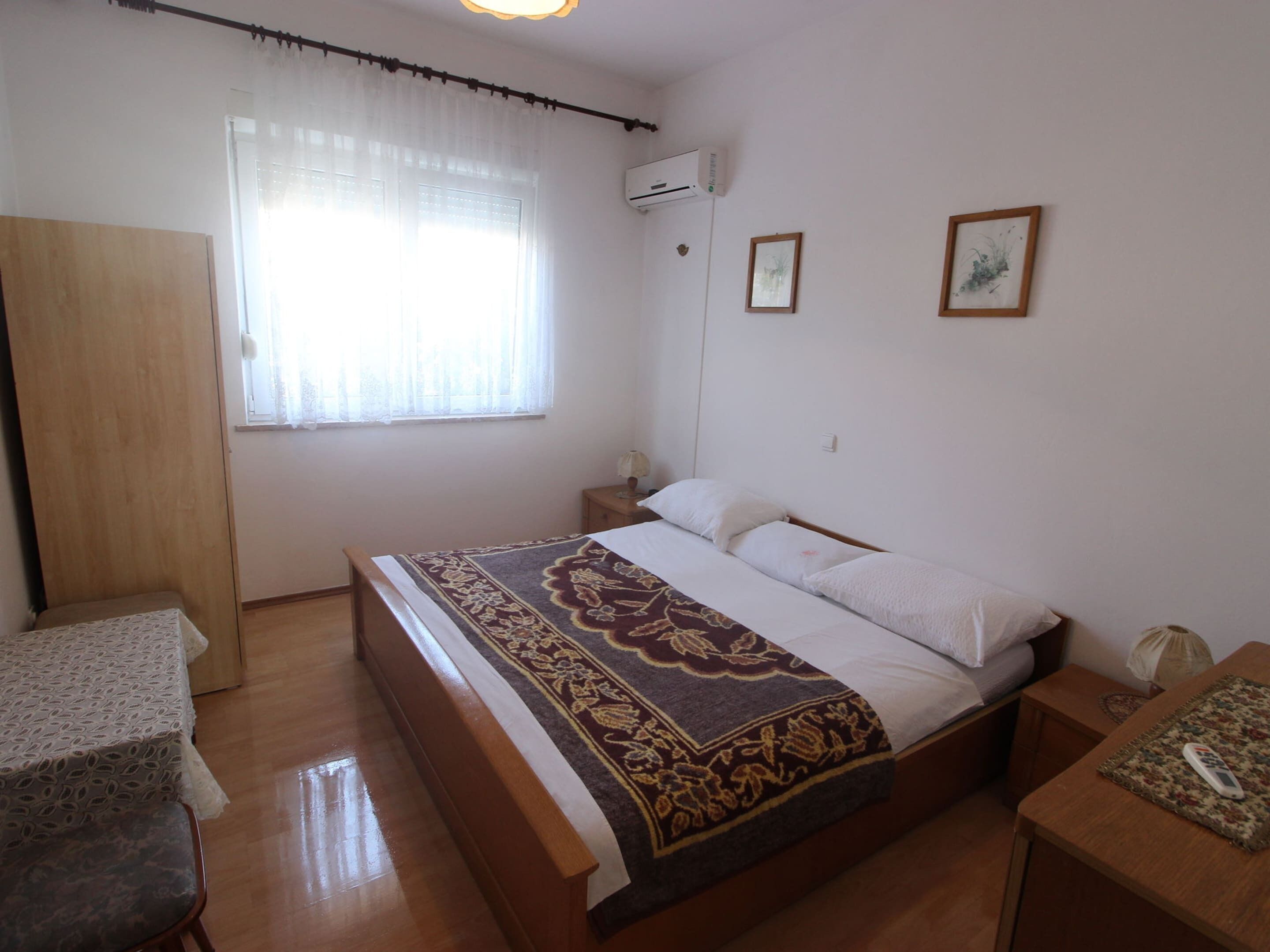 A bedroom with a bed, a nightstand, a wooden wardrobe, a window with sheer curtains, and two framed pictures on the wall.