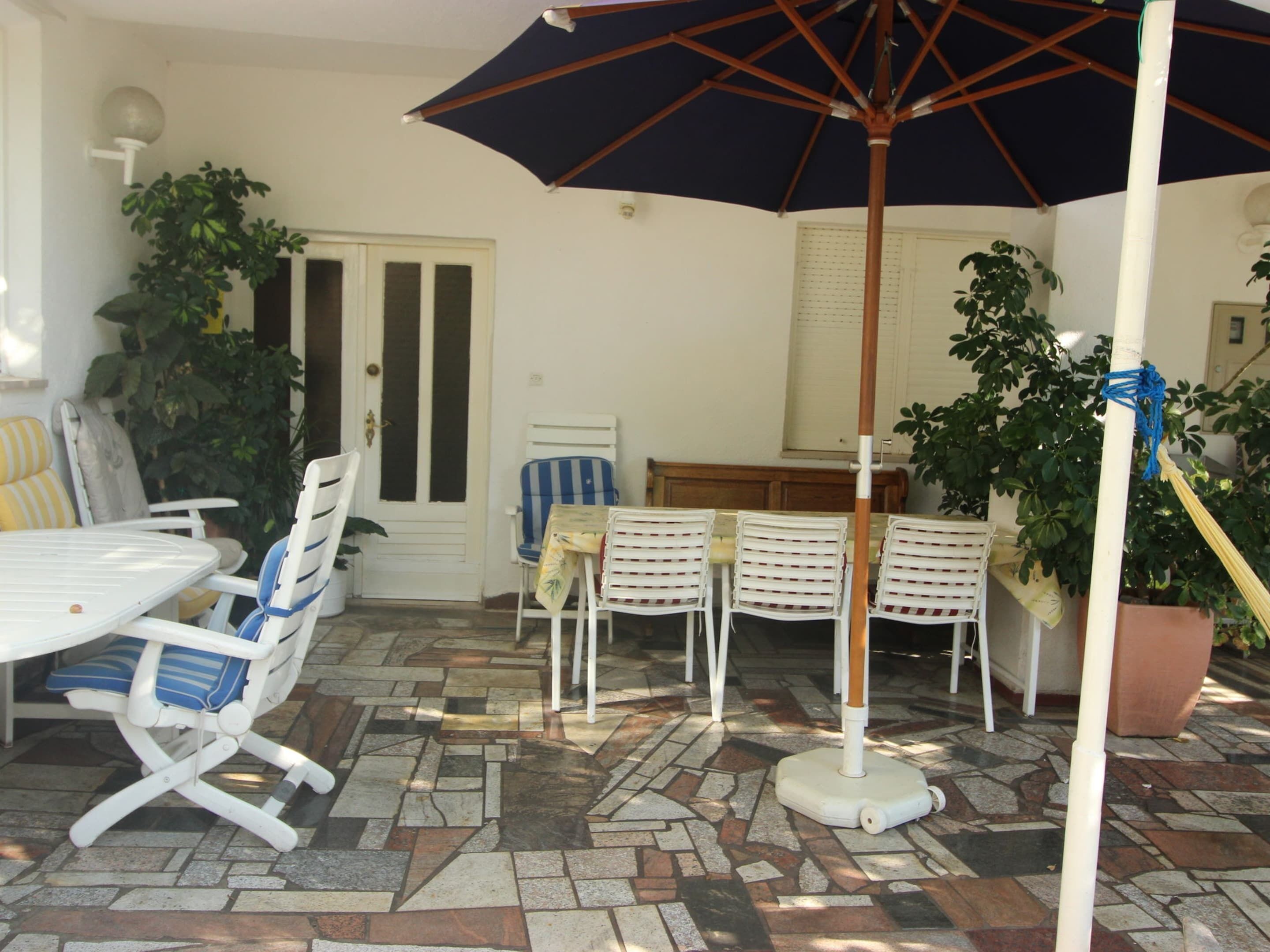 Un patio al aire libre con una mesa y sillas blancas, una sombrilla azul y plantas en macetas.