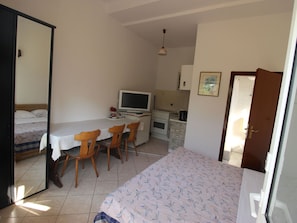 Quarto pequeno e compacto com cama, mesa de jantar com cadeiras, cozinha compacta e televisão.