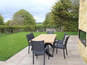 A patio with a wooden table and black wicker chairs, surrounded by a stone wall and a grassy lawn. - Bemelen Holiday Home for 6 (Bemelen)