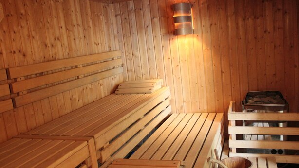A wooden sauna with benches and a bench seat, a wooden bucket, and a sauna heater. - Winenne Home With Hot Tub (Beauraing)