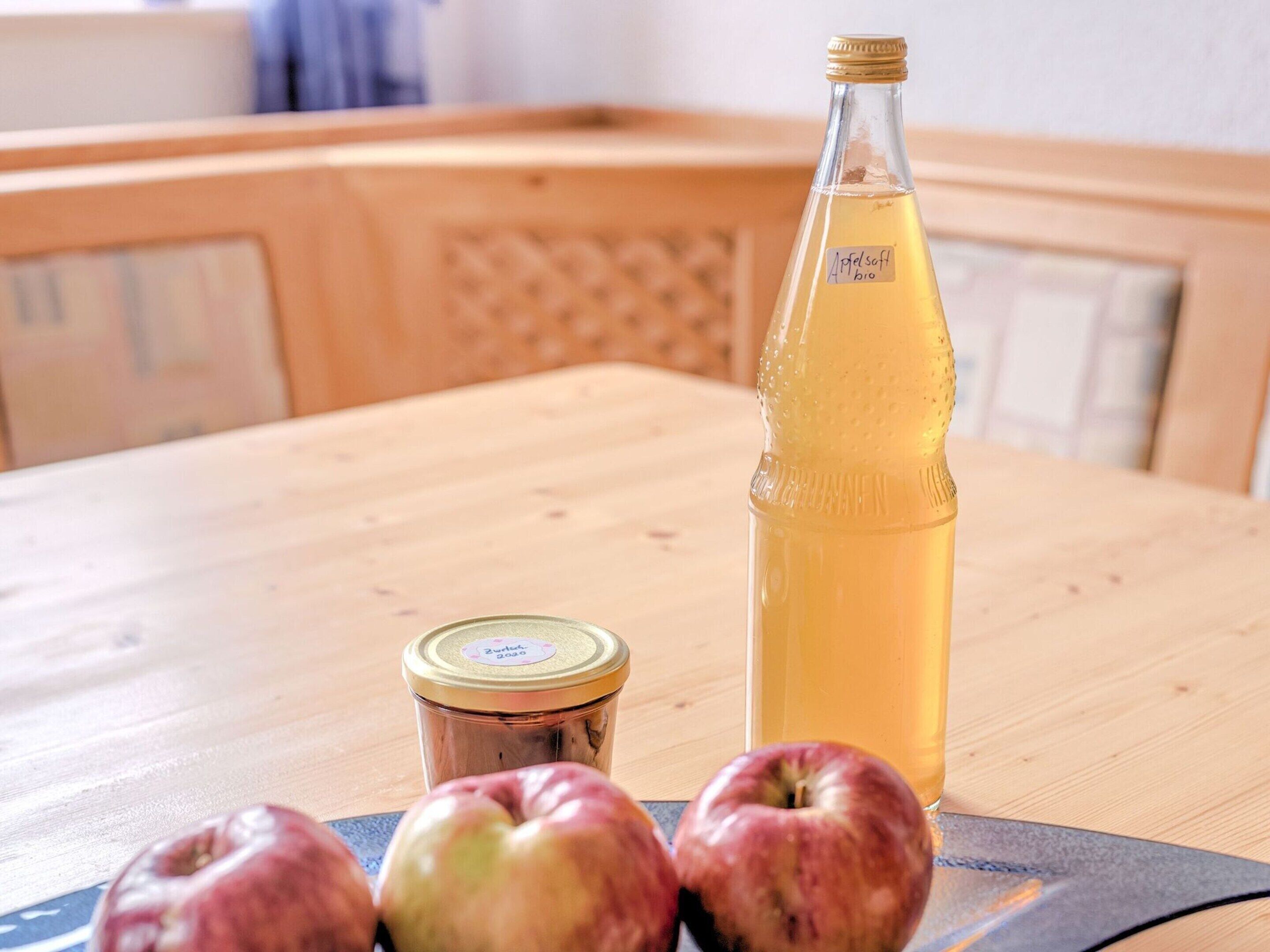 A wooden table with a bottle of apple cider, a jar, and three apples.