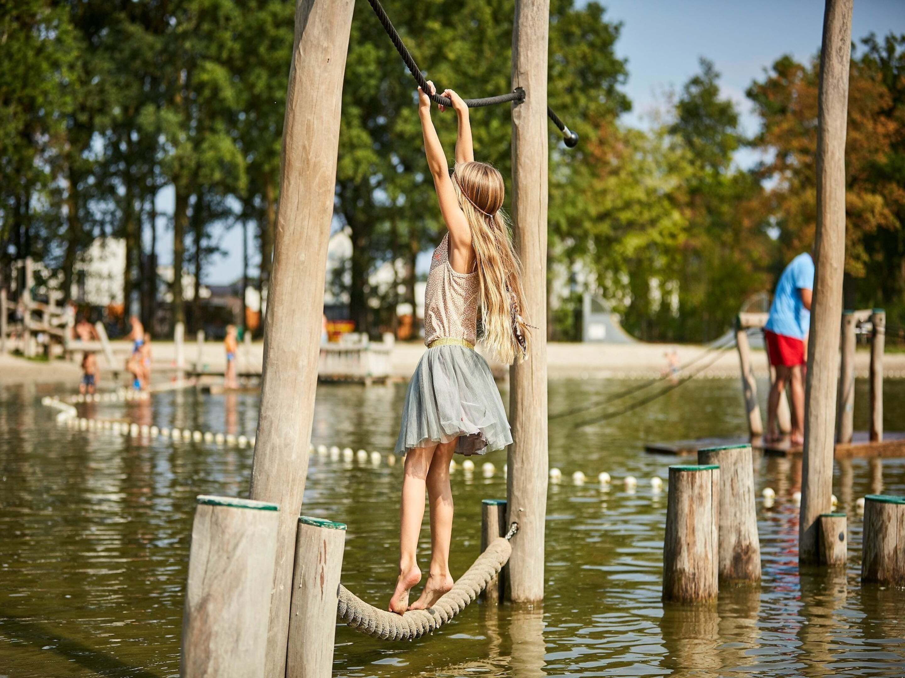 A person on a rope bridge over water, surrounded by wooden posts and trees.