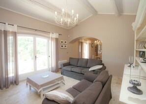 A living room with a sofa, a table, a chandelier, and a view through a glass door. - Villa in Provence With Infinity Pool (Apt)