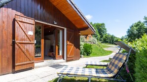 Eine Holzhütte mit großer offener Tür, einer überdachten Terrasse mit Liegestuhl und Blick auf eine grüne Landschaft.