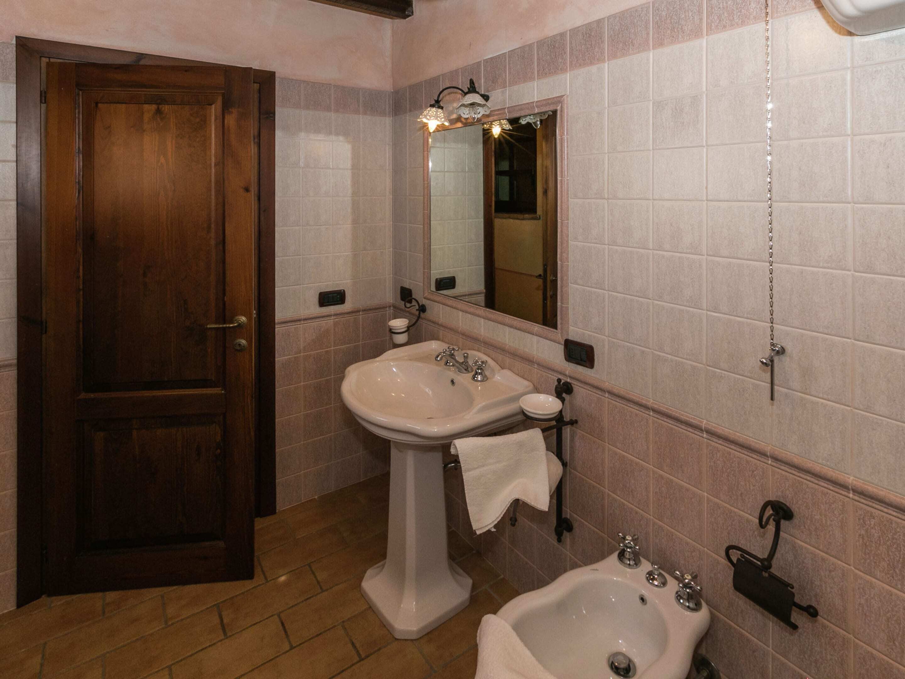 A bathroom with a wooden door, a pedestal sink, a bidet, and a mirror with lights.