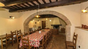 A dining area with a long table covered in a floral tablecloth, surrounded by wooden chairs. An arched brick wall separates the dining area from a fireplace. - Luxury, Modern Apartment With Pool and Stunning Views (Narni)