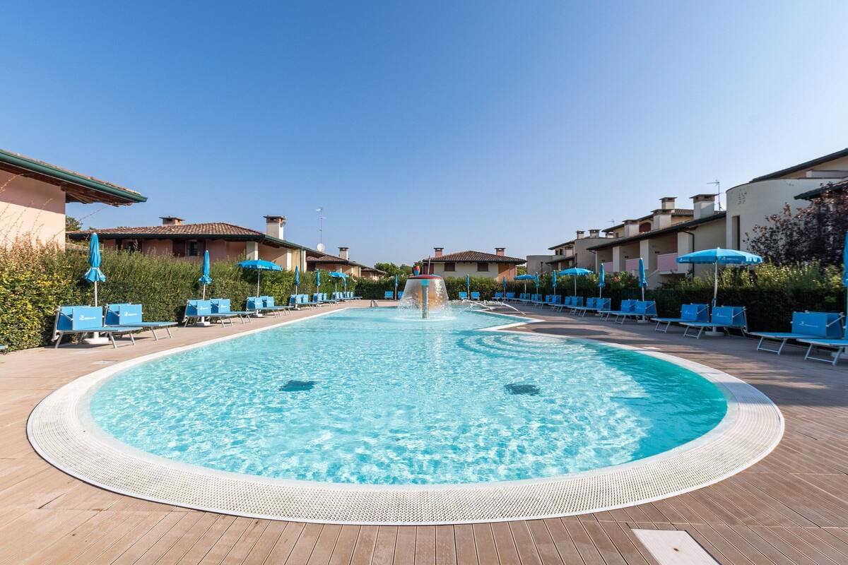 A swimming pool with a central water feature, surrounded by lounge chairs and umbrellas, with buildings in the background.