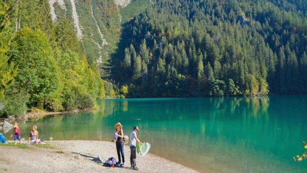 Ein See, umgeben von bewaldeten Bergen, mit Personen am Ufer.