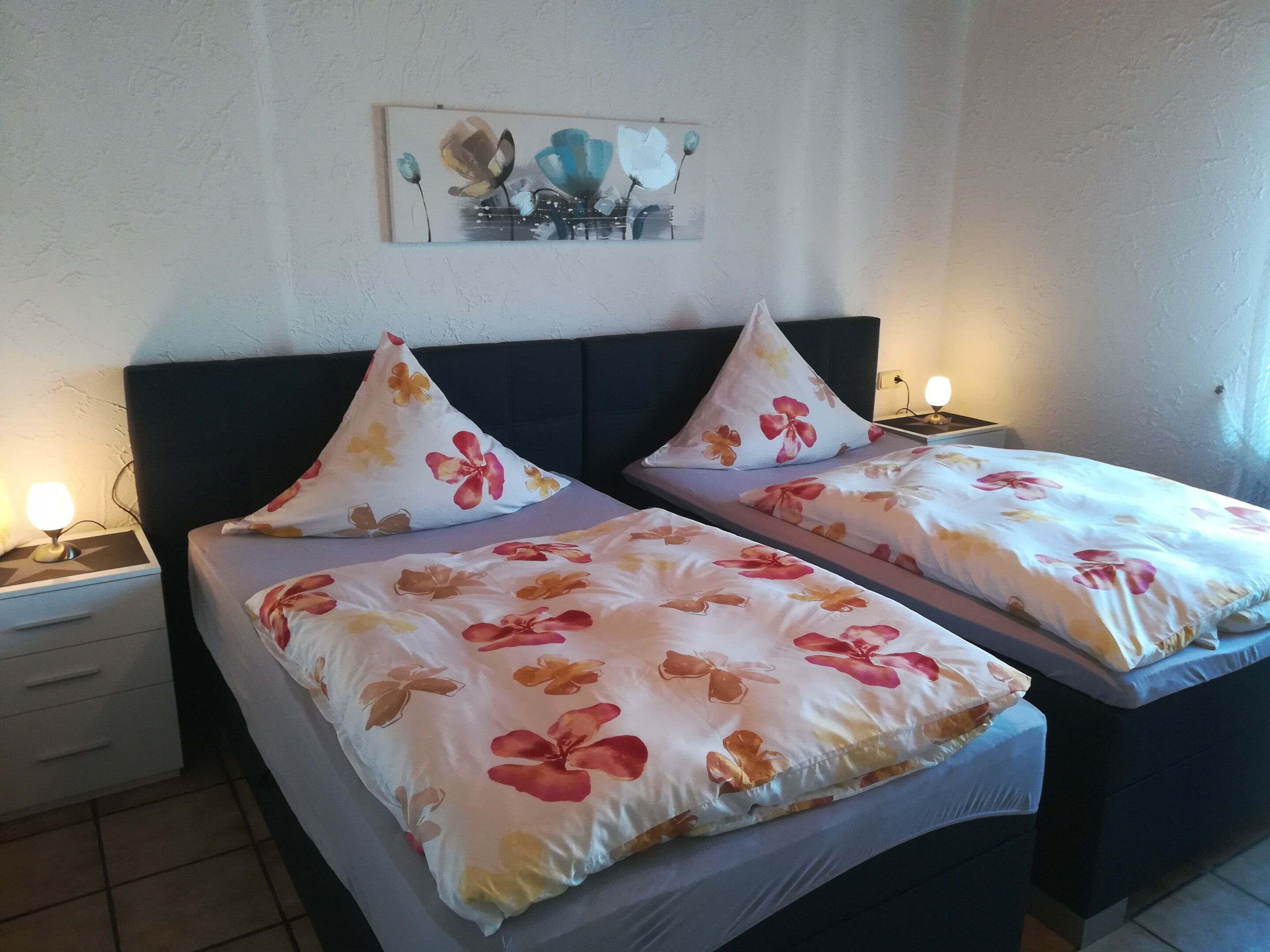 Une chambre d’hôtel avec deux lits, chacun agrémenté de draps et d’oreillers à motifs floraux. On y trouve une table de chevet avec une lampe, et une œuvre d’art est accrochée au mur au-dessus des lits.