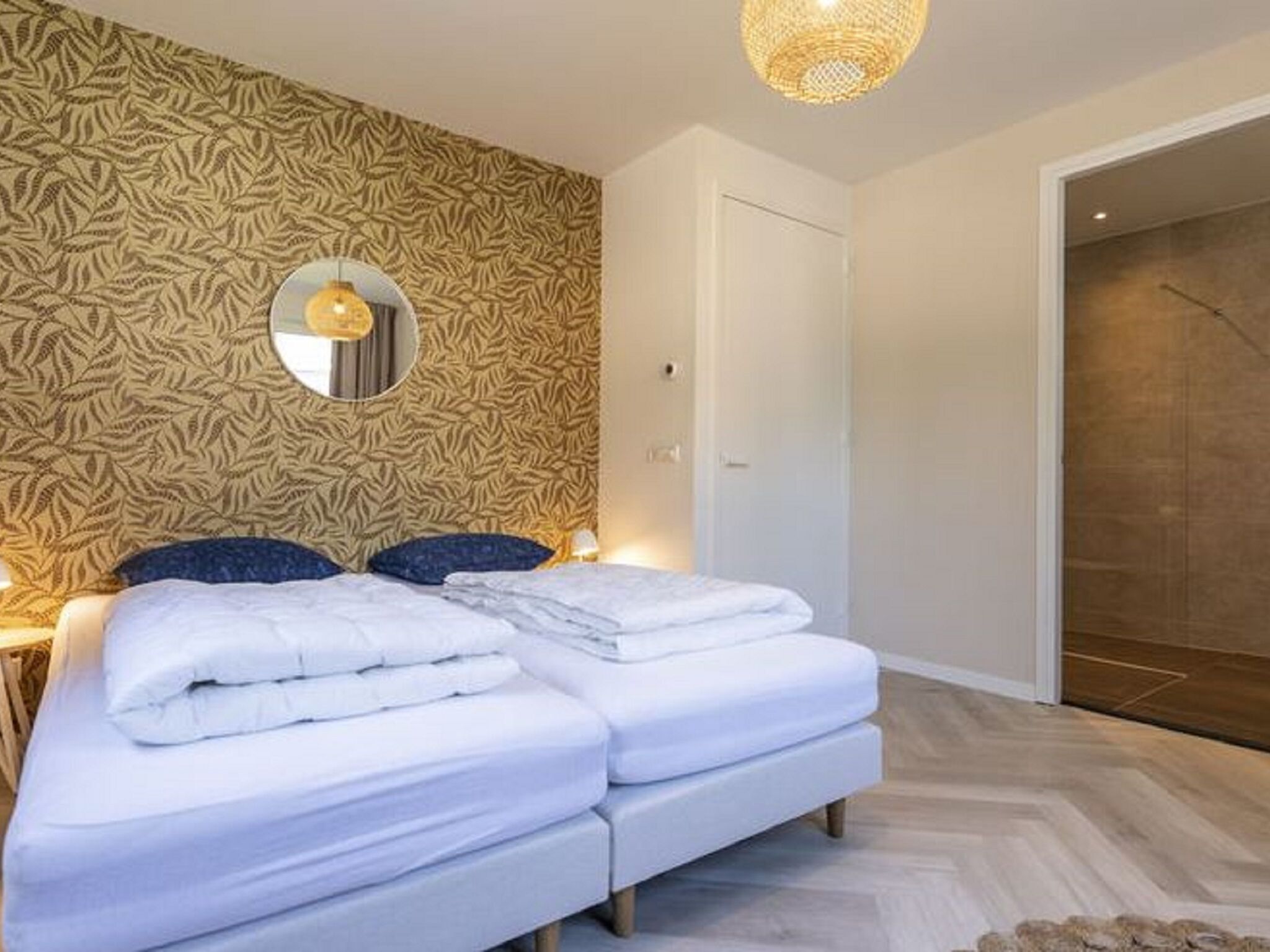 A bedroom with a patterned wallpaper, a bed with white bedding, a round mirror, and a hanging light fixture.
