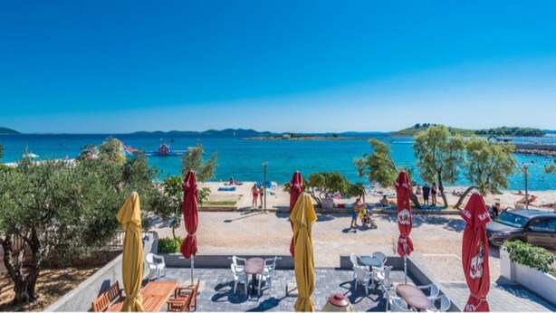 Eine Terrasse mit Sitzgelegenheiten und Sonnenschirmen mit Blick auf einen Strand, an dem sich Personen und Boote befinden.