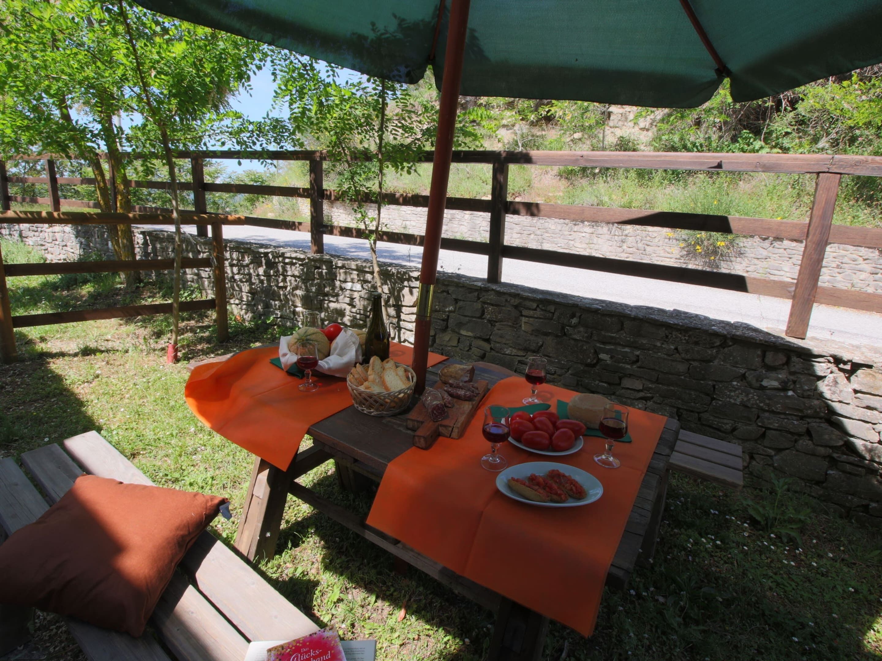 An outdoor dining area with a table set for two, covered by a green umbrella, surrounded by a stone wall and wooden fence.