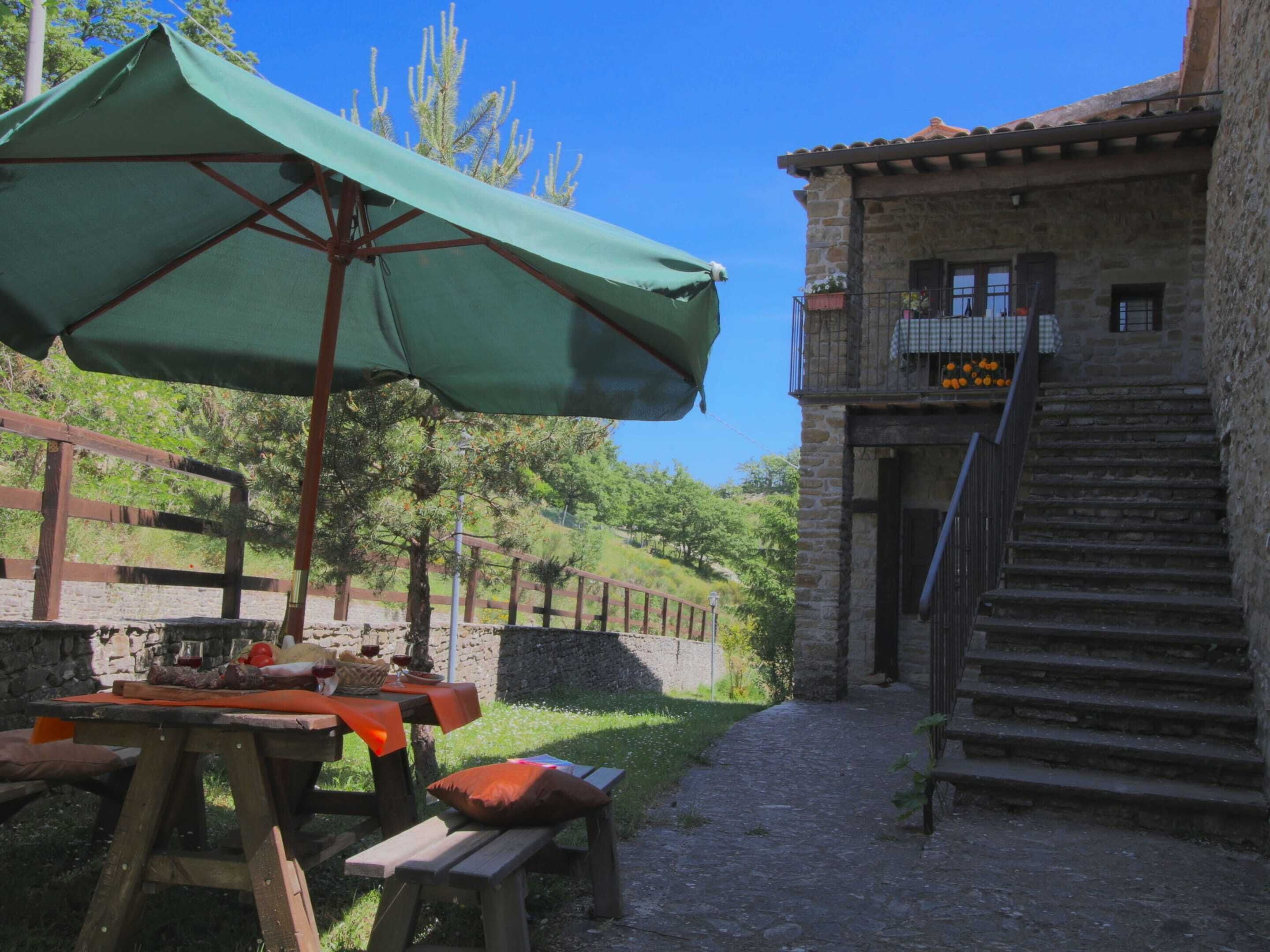 A rustic outdoor dining area with a table and benches, a green umbrella, and a stone building with a staircase.