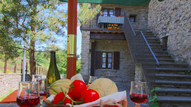 A rustic outdoor dining setup with a stone building in the background, a wooden table with food and wine, and a staircase leading to a balcony.