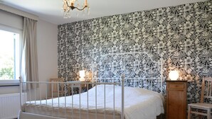 A bedroom with a white metal bed frame, floral wallpaper, a wooden cabinet, and a chandelier.