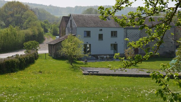 A large, light-colored house with a sloped roof, surrounded by a well-maintained lawn and a small garden area.