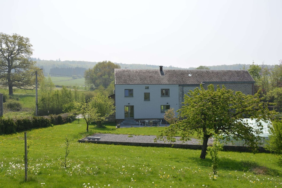 Ein zweigeschossiges Haus mit Ziegeldach, umgeben von einer Wiese mit Löwenzahn und einigen Bäumen.