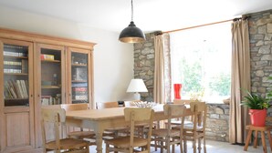 A dining area with a wooden table and chairs, a lamp, a bookshelf, and a stone wall.