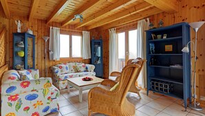 Ein gemütlicher Raum mit Holzwänden und -decke, einem blumigen Sofa, Korbstühlen, einem kleinen Tisch und einem Bücherregal.