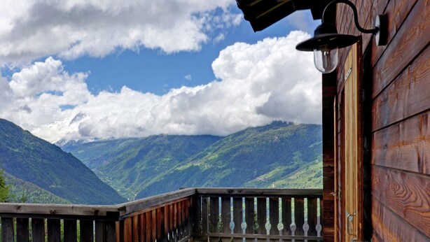 Ein Holzbalkon mit Metallgeländer, der auf eine gebirgige Landschaft unter leicht bewölktem Himmel blickt.