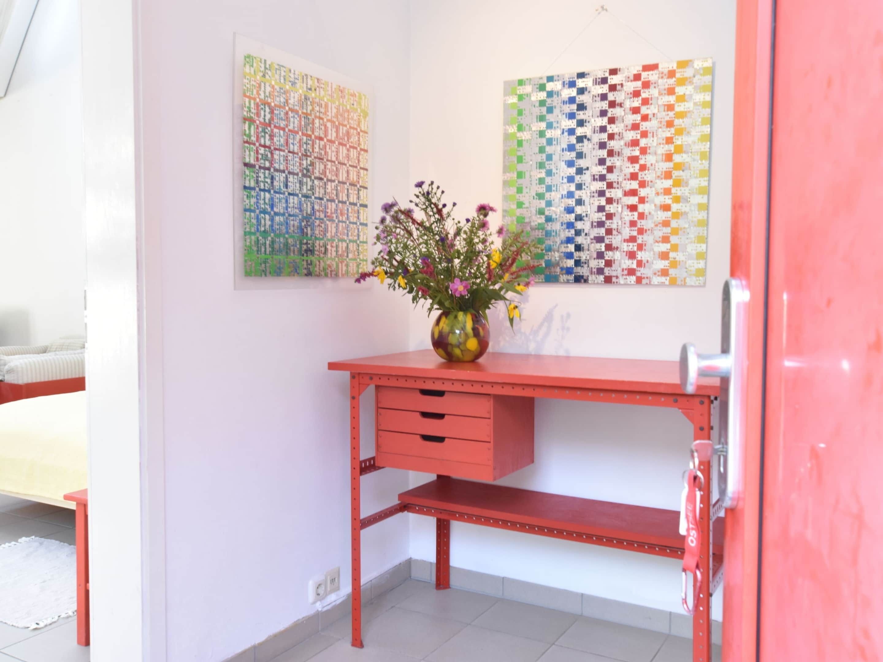 Una stanza con una scrivania rossa, un vaso con fiori e due quadri astratti colorati appesi al muro.
