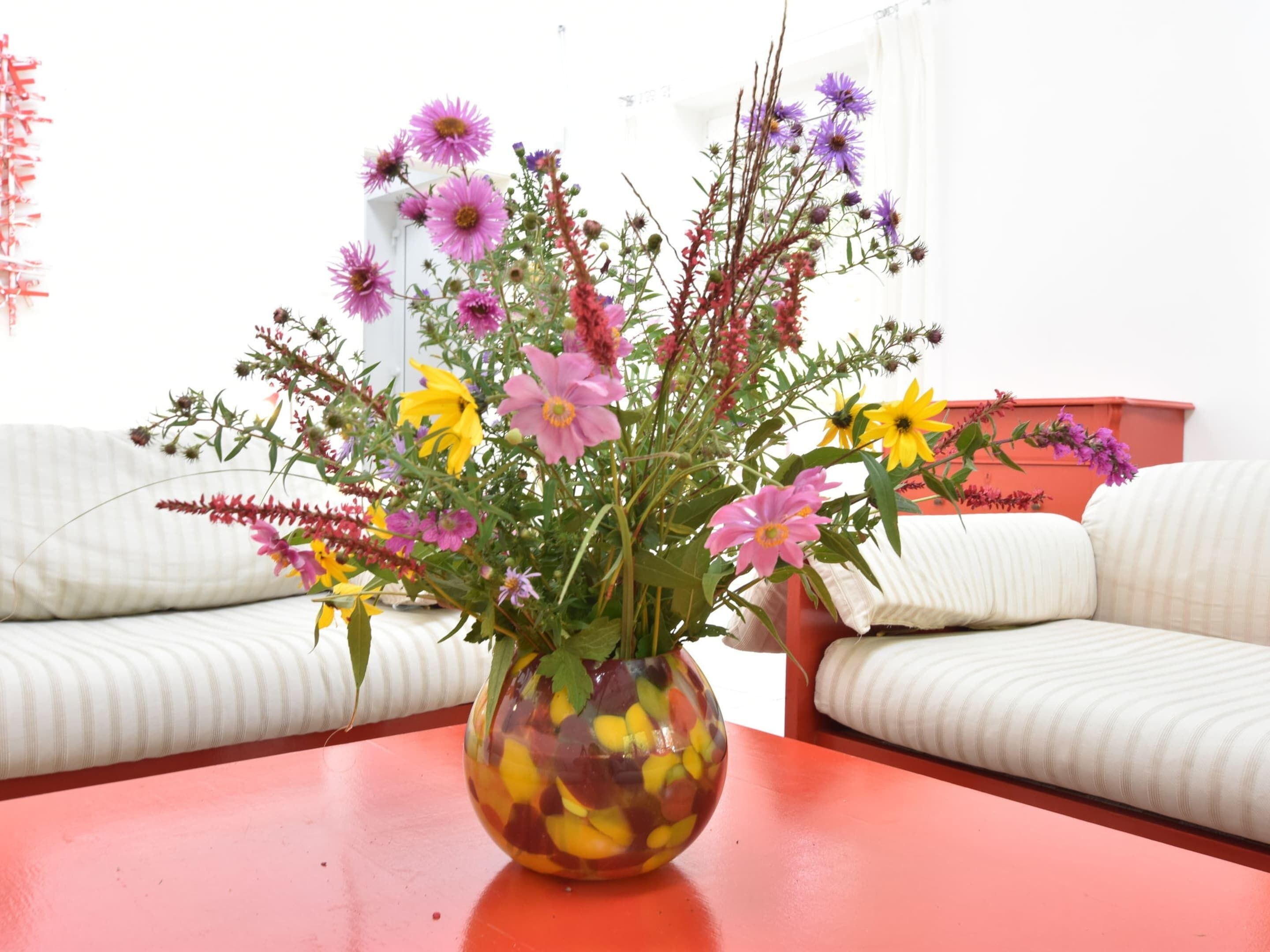 A room with a red table, a white sofa, and a vase with flowers.
