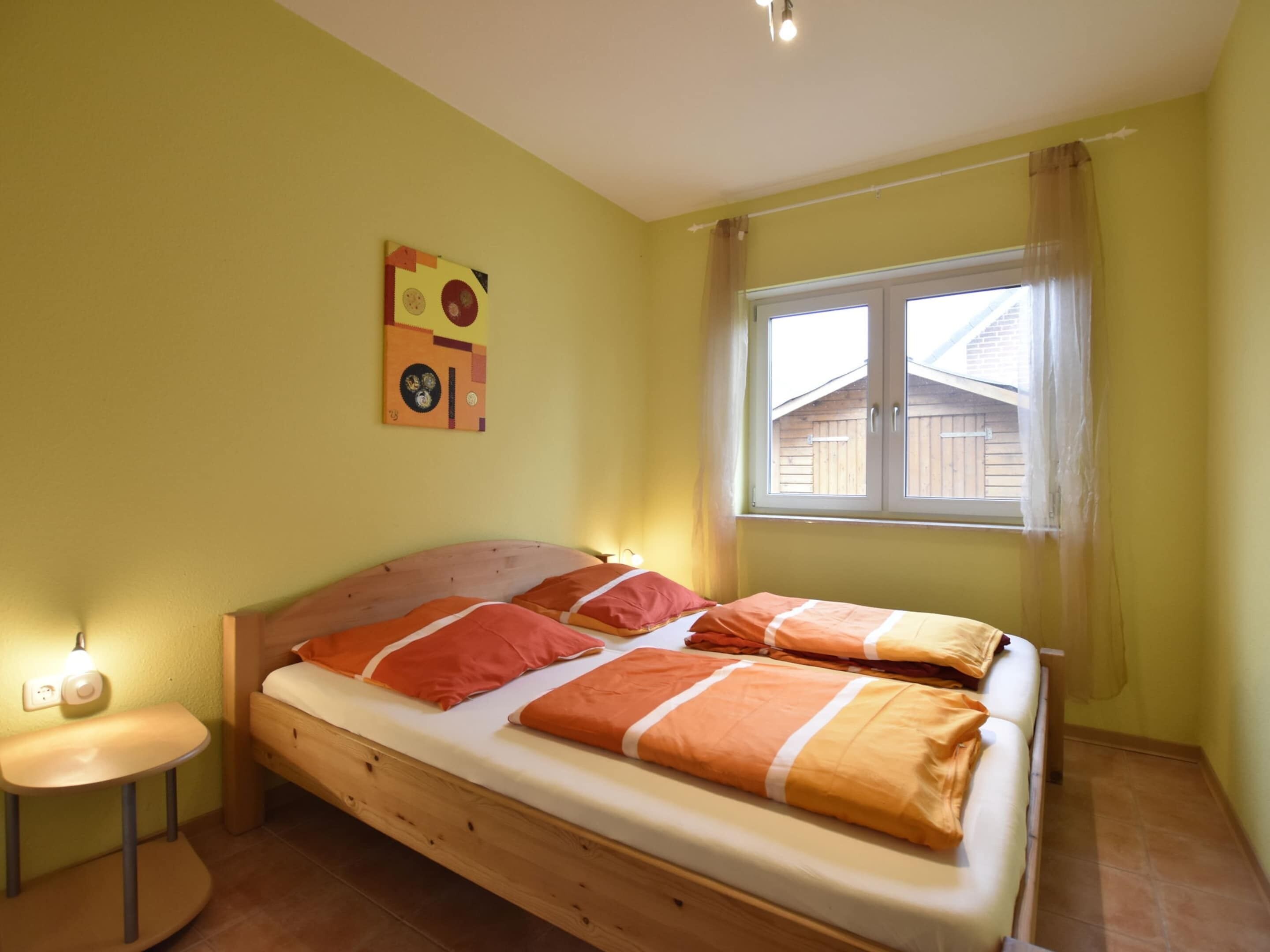 A single bed with orange and white bedding, a small bedside table, a wall clock, and a window with curtains.