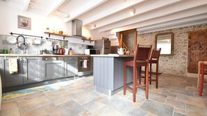 Une cuisine avec des meubles de couleur grise, un îlot central, des chaises en bois et un mur en pierre.