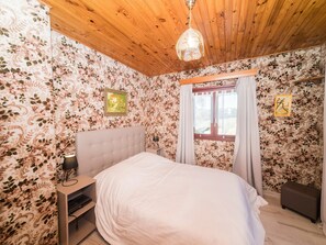 A bedroom with a wooden ceiling, floral wallpaper, a bed, a nightstand, a lamp, a window with curtains, and a small stool. - House in Vosges Near Cascades du Gerhard (Girmont-Val-d'Ajol)