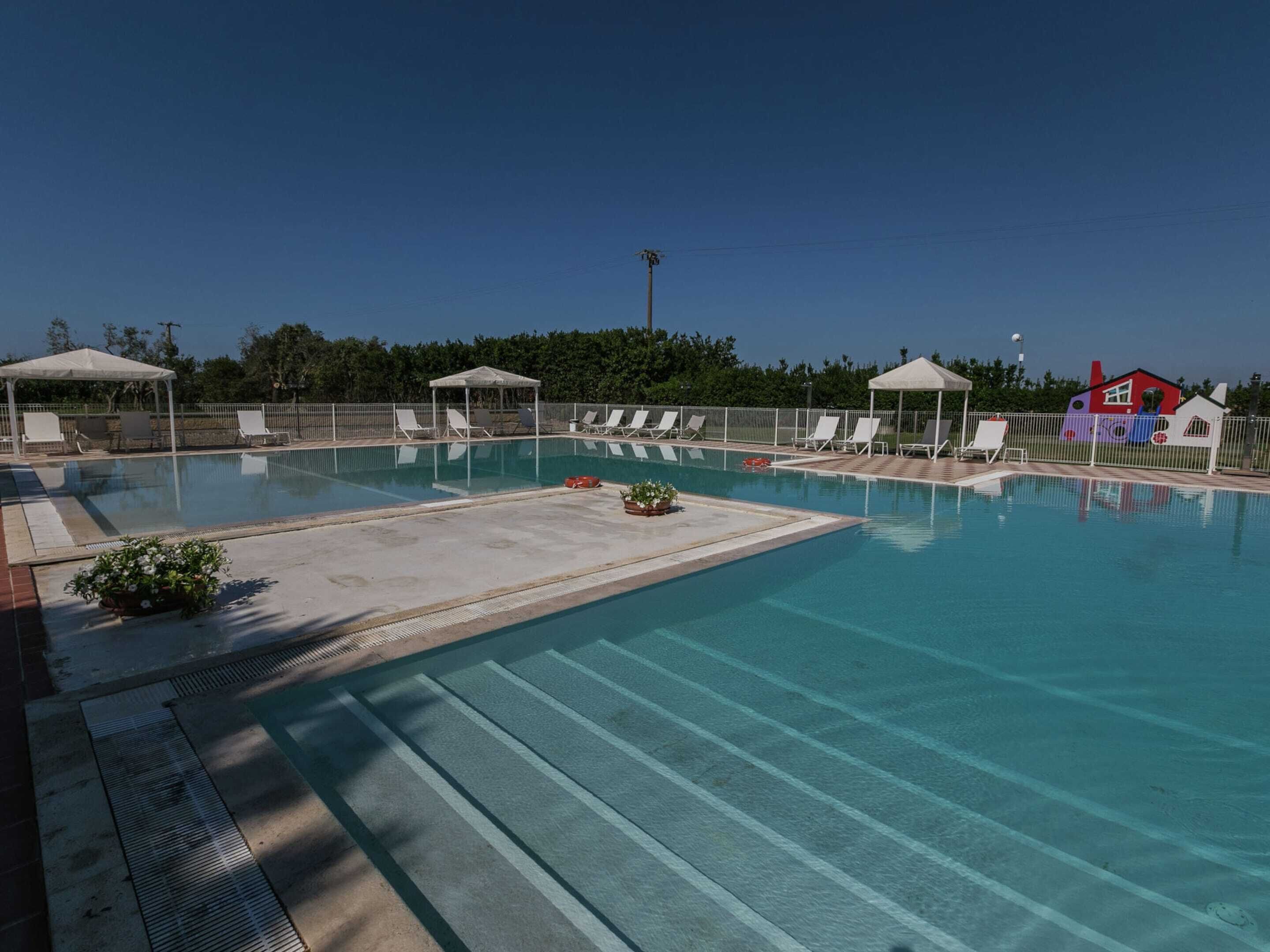 Una piscina con lettini e ombrelloni, circondata da una recinzione e vegetazione.